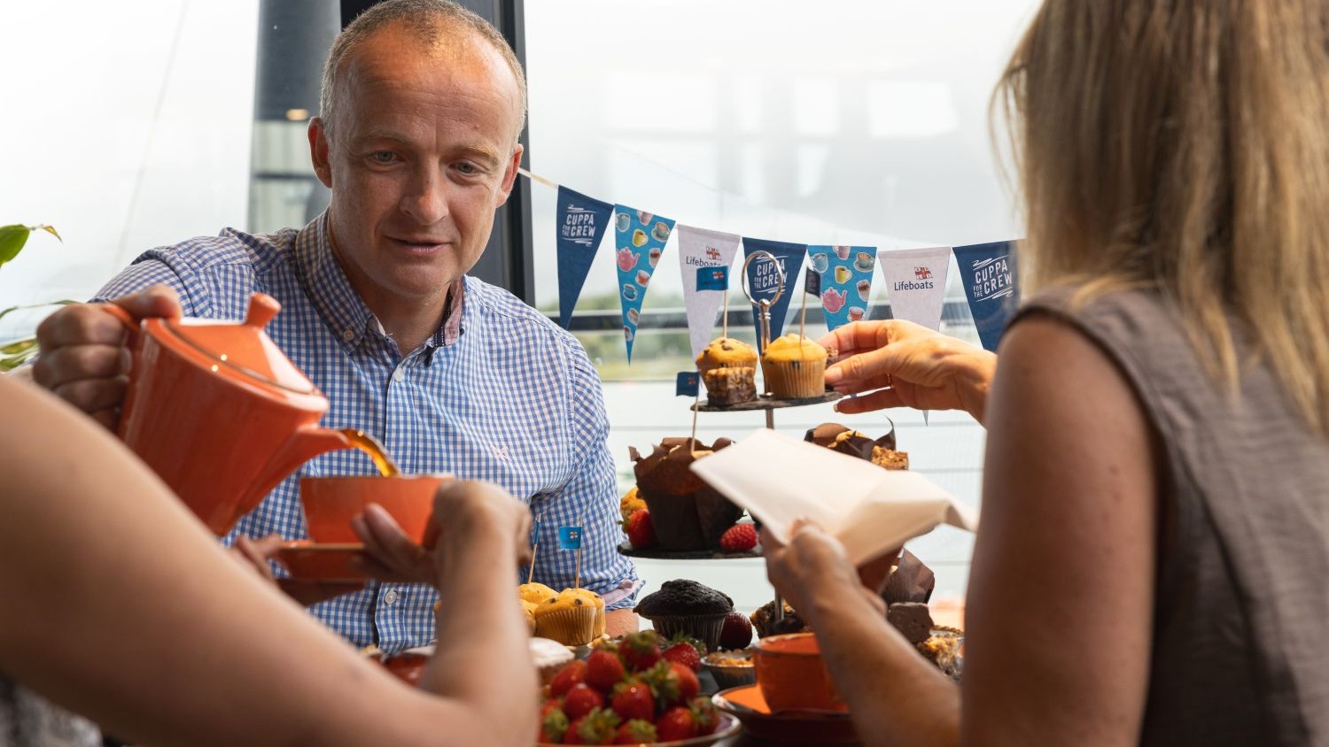People gathered around a table at a charity tea event, pouring tea and serving cakes and muffins. Decorative bunting featuring RNLI Lifeboats and “Cuppa for the Crew” branding is displayed in the background, with plates of strawberries and baked treats on the table.